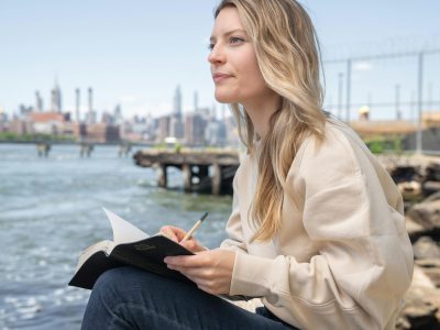 Mujer rubia escribiendo en un cuaderno mientras reflexiona junto al agua